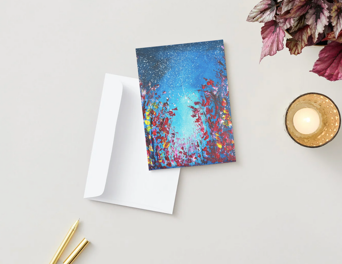A6 Starnight greeting card with vibrant blue and red painted forest design, paired with a white envelope on a desk.