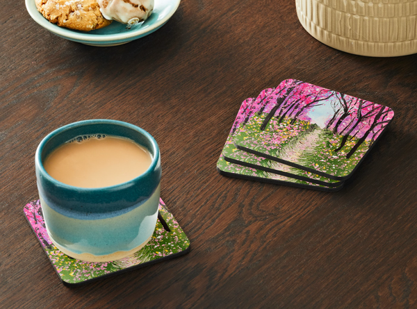 Glossy 9.5cm hardboard coaster featuring a vibrant pink cherry tree walkway painting, set on a dark wooden table.