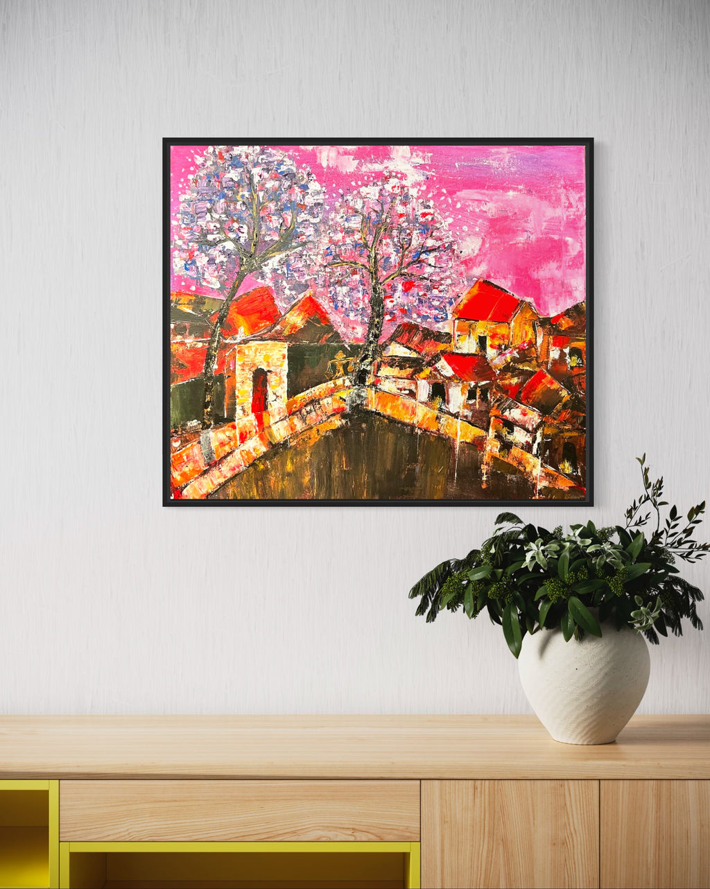 Framed abstract cityscape painting with orange houses, bridge curve, and pink sky background.