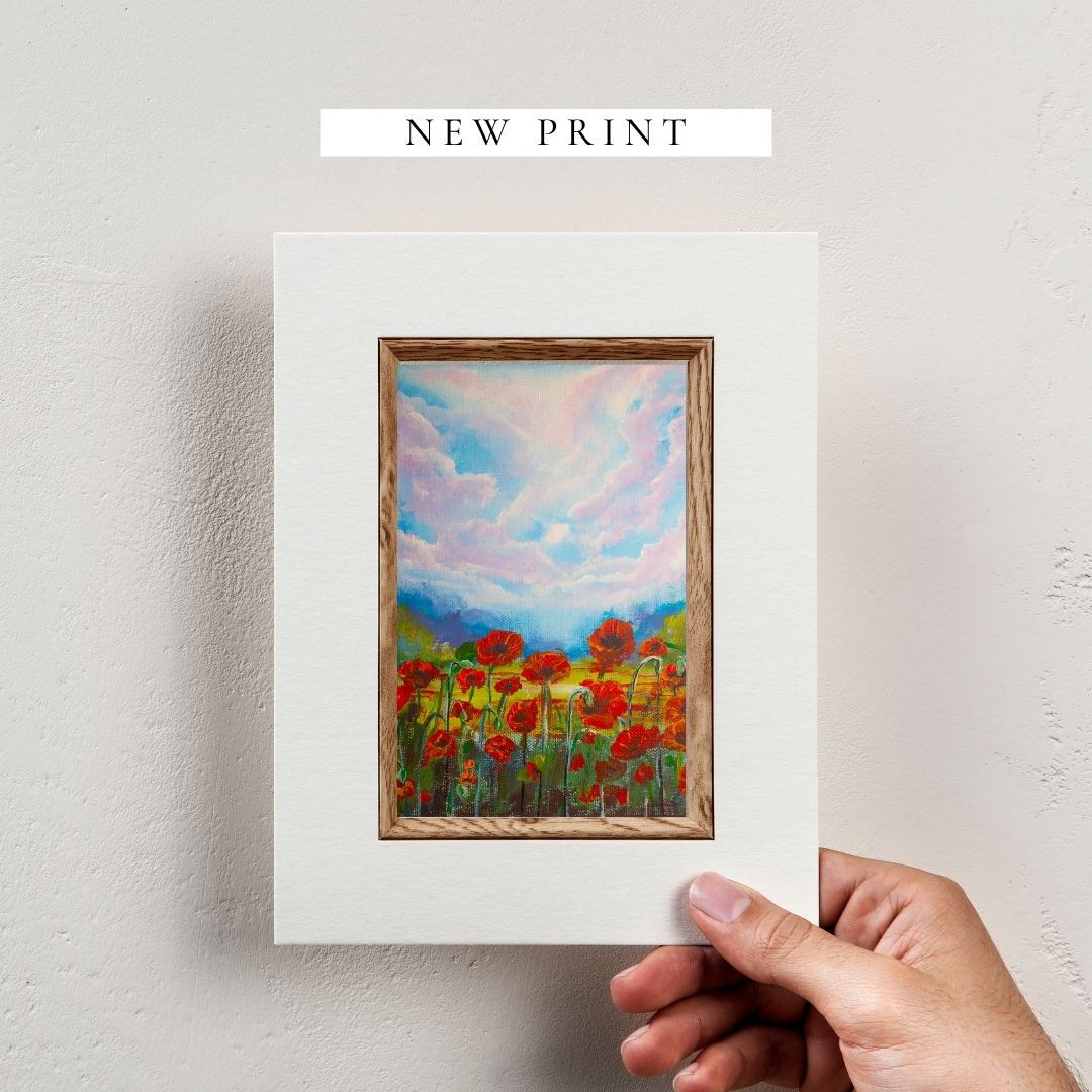 Hand holding a framed wildflower artwork, red flowers under blue-pink cloudy sky