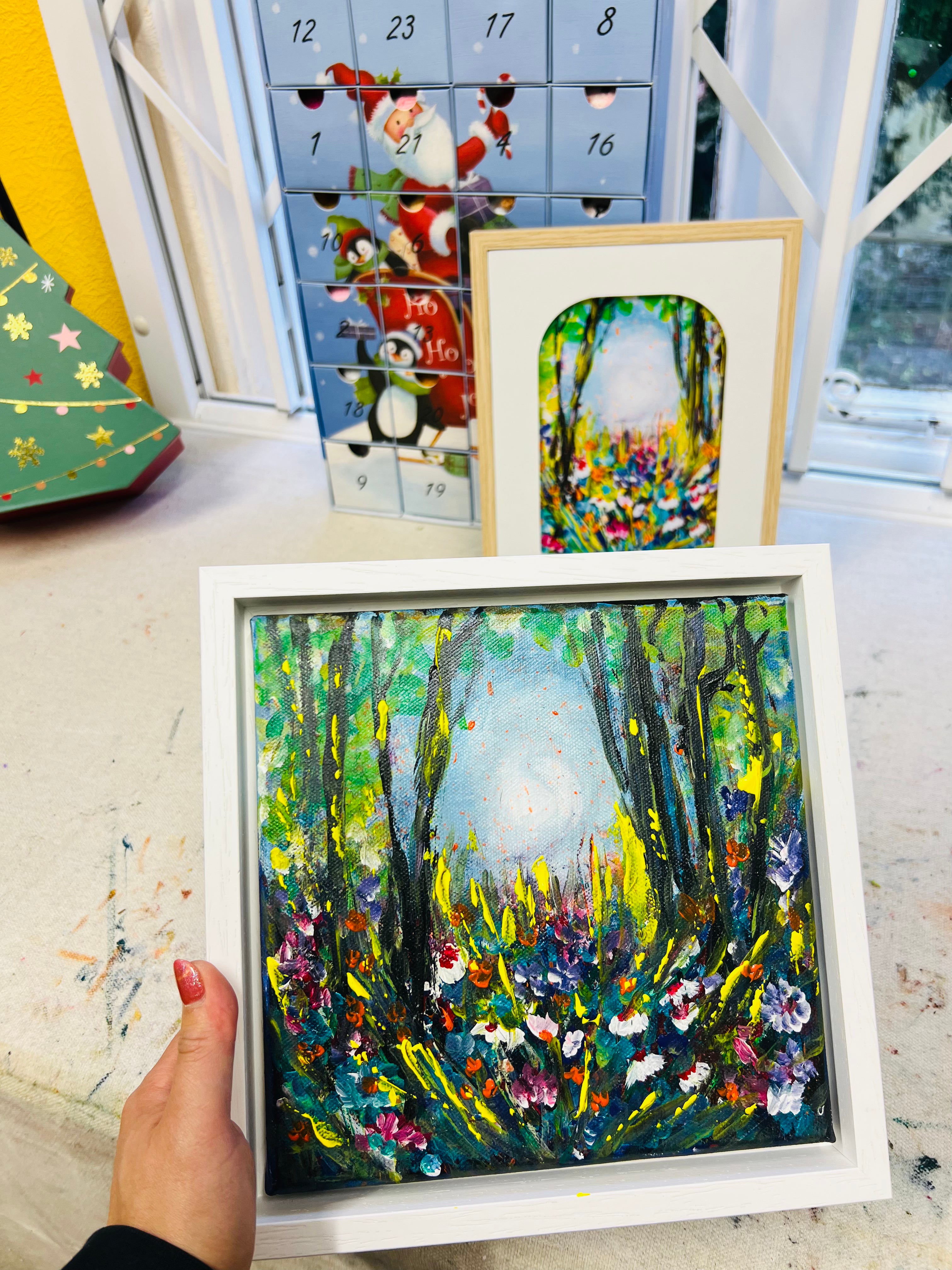 Hand holding a square, white-framed art print featuring a vibrant, textured oil painting of a colorful forest meadow.