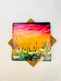 Square ceramic coaster showing a vibrant sunset sky over tall grass and flowers