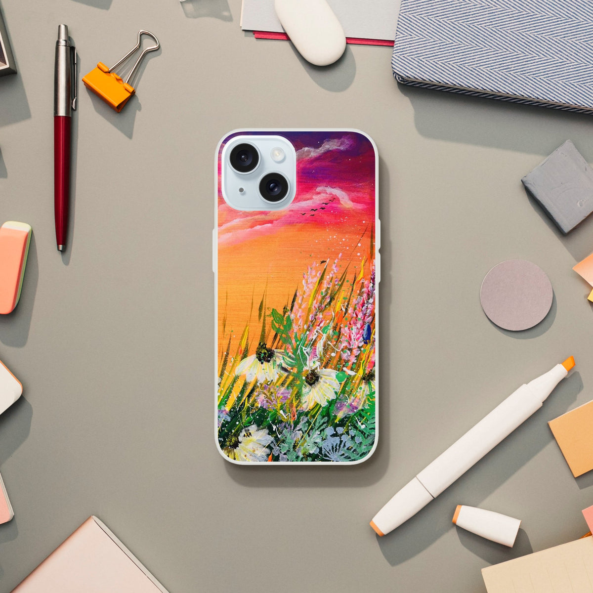 Flexible phone case featuring a vibrant sunset landscape painting with wildflowers on a desk with office supplies.